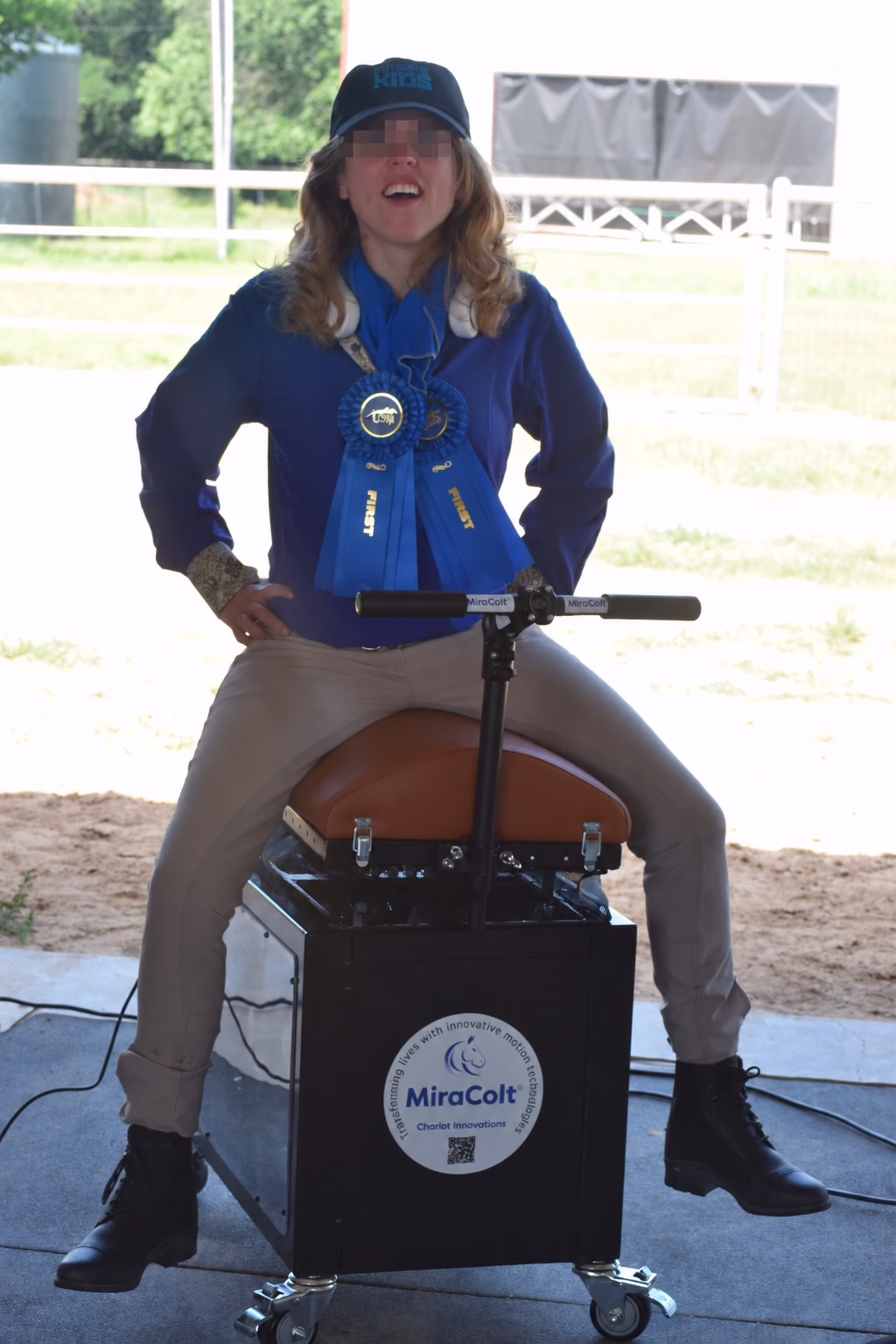 Participant wearing blue riding ribbons seated on the MiraColt at an equine program.